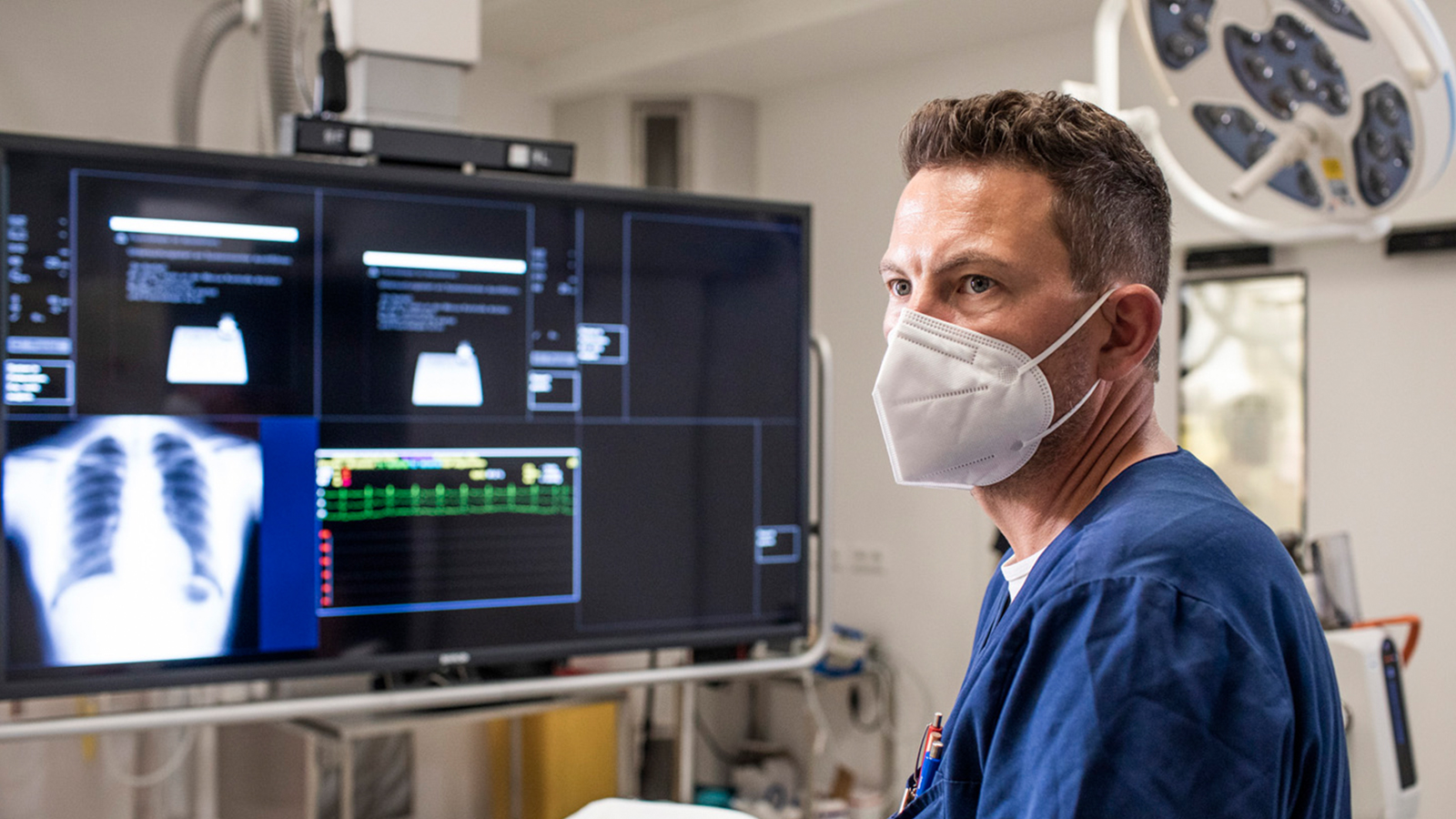 Ein Arzt oder Pfleger aus der Radiologie steht an einem Patientenbett, im Hintergrund ein riesiger Bildschirm mit einer abgebildeter Lunge.