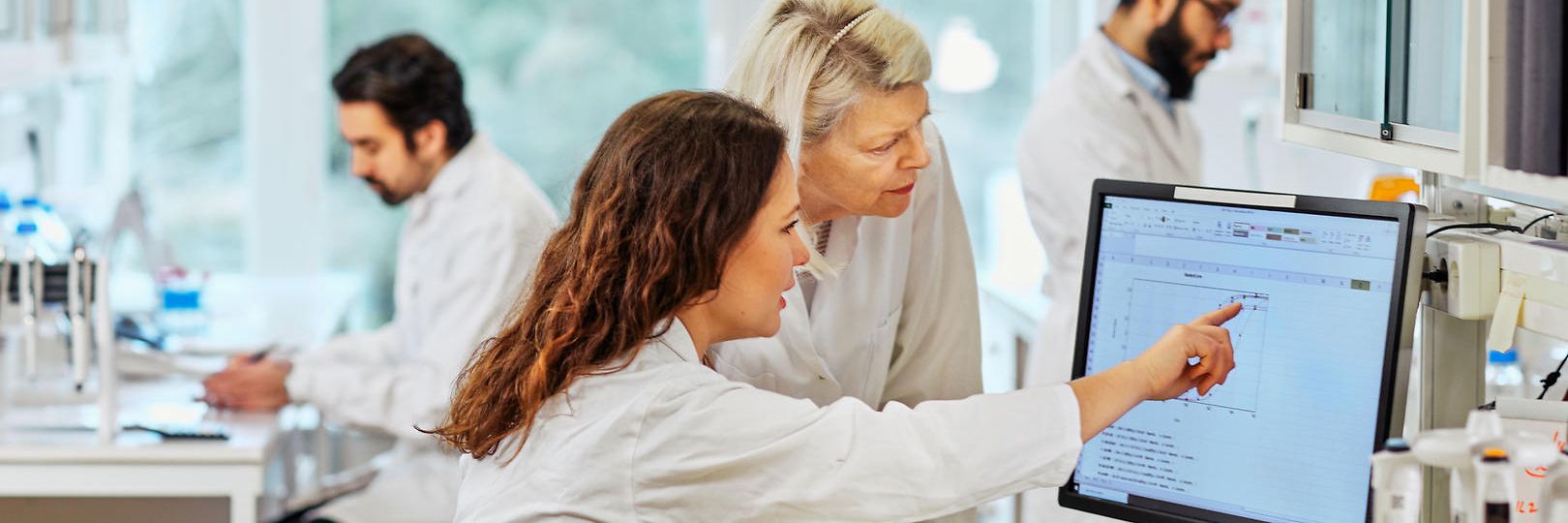 Eine Frau zeigt Ihrer Laborkollegin etwas auf dem Bildschirm. Zwei Kollegen gehen ebenfalls ihrer Arbeit nach.