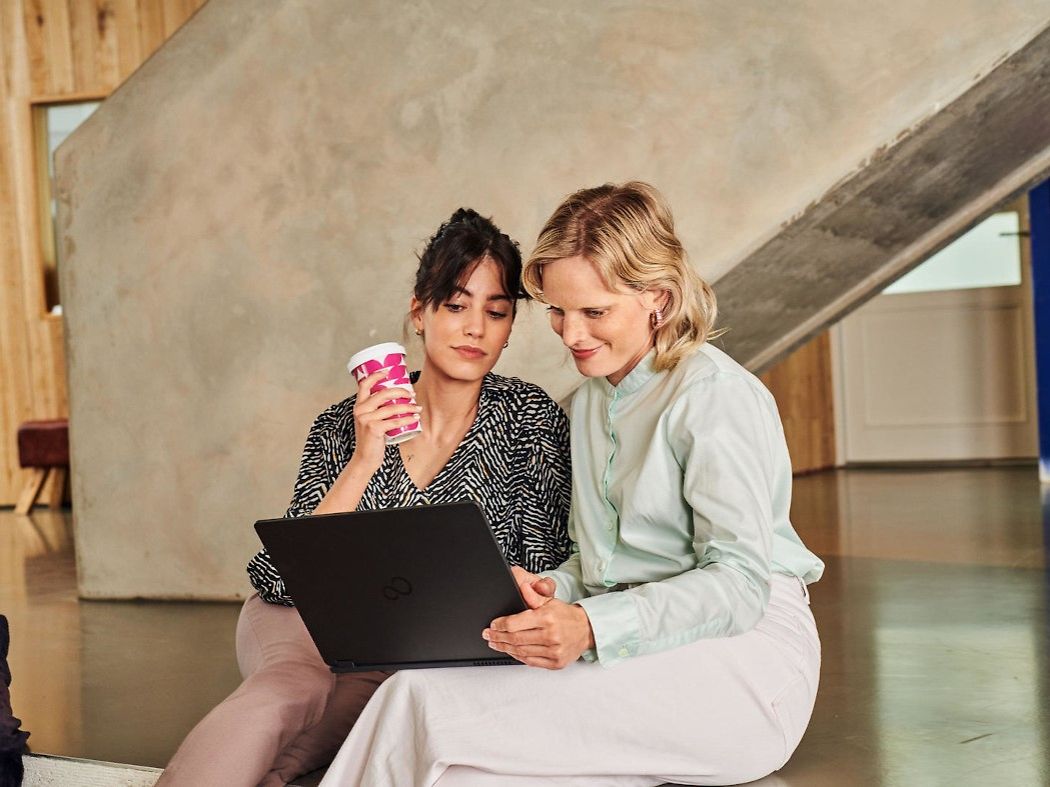 Frau mit Kaffeebecher schaut gemeinsam mit einer anderen Frau auf einen Laptop