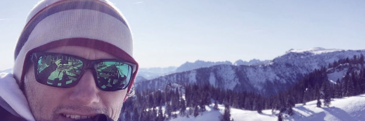 Mann mit Sonnenbrille und Mütze vor verschneiter Bergkulisse