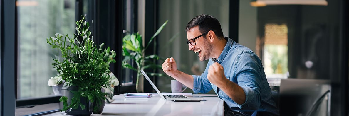 Mann mit hochgekrempelten Ärmeln und Brille hebt freudig die Fäuste während er auf den Laptop auf seinem Schreibtisch blickt