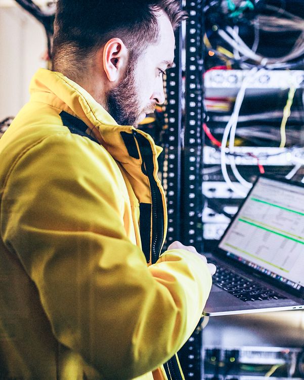 Ein Mann steht vor einem Serverschrank und schaut auf seinen Laptop.
