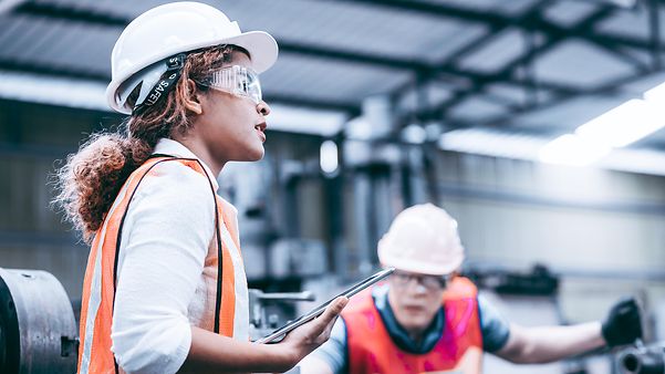 Eine Frau steht mit Helm und Warnweste und Tablet in der Hand in einer Halle.