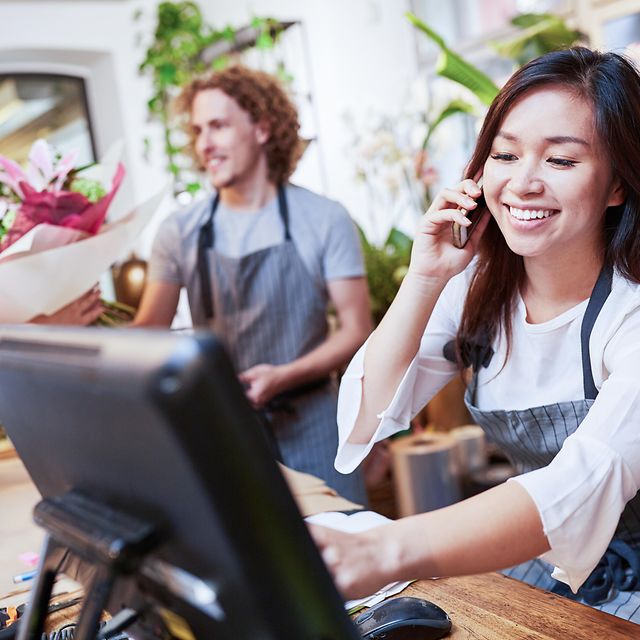 A young woman and a young man serve customers in a flower shop with a digital cash register.