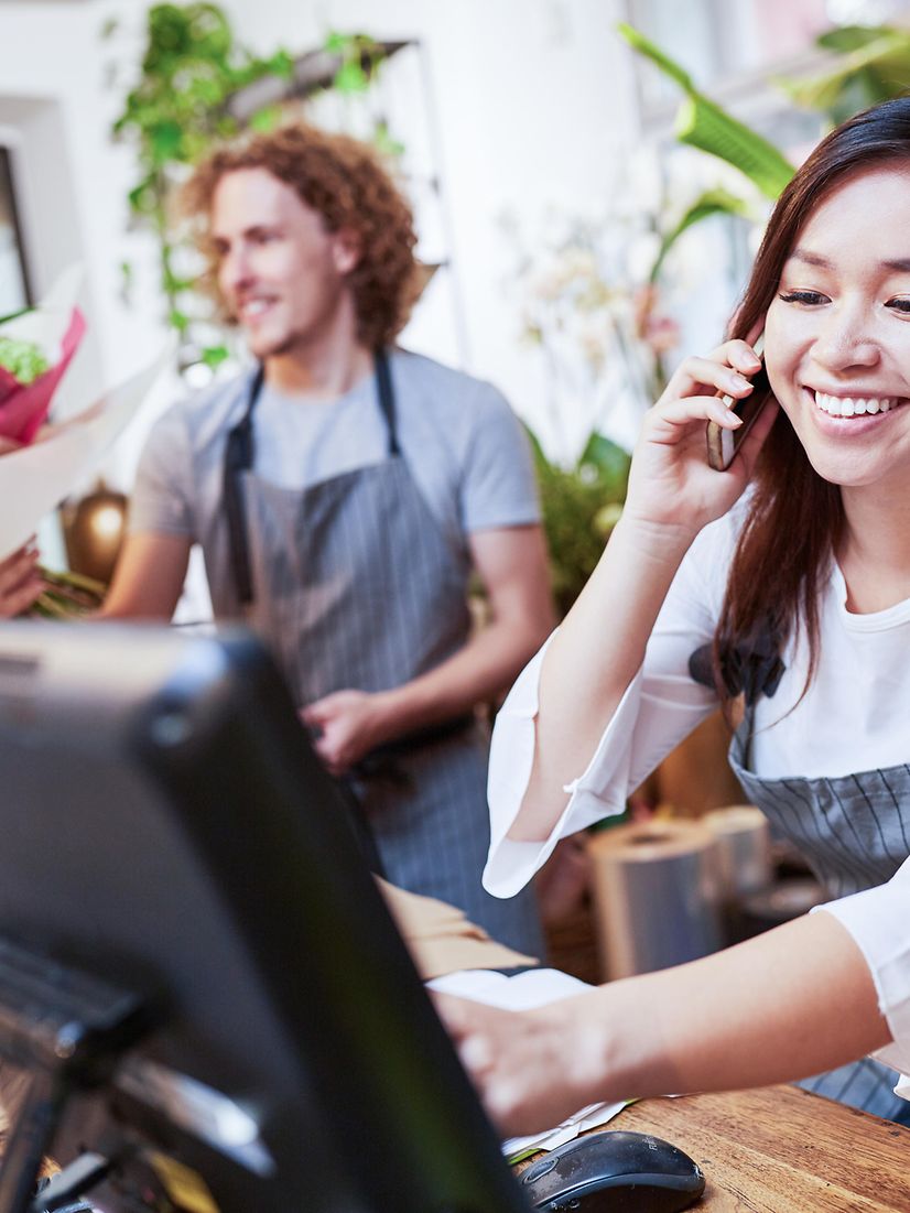 A young woman and a young man serve customers in a flower shop with a digital cash register.