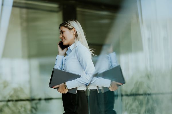 A woman in business attire is on the phone and carrying a notebook under her arm.