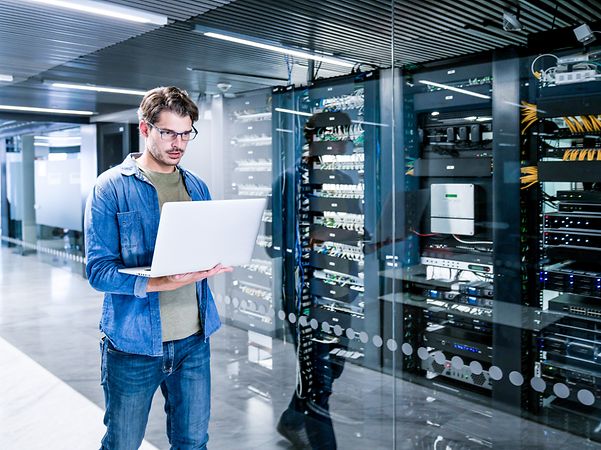 An employee is standing in the server room with a notebook.