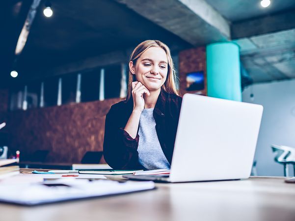 A woman sits at a table with documents in a modern office and looks at the screen.