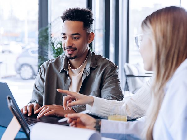 Two people are sitting together at a desk at their computers and talking to each other.
