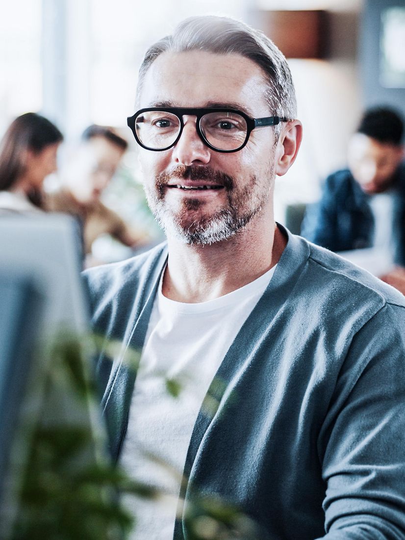 Man in office at computer, team meeting in background