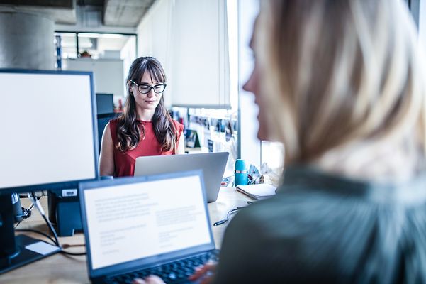Looking over the shoulder of one colleague to another colleague who has her notebook with Zoom X Rooms in front of her.