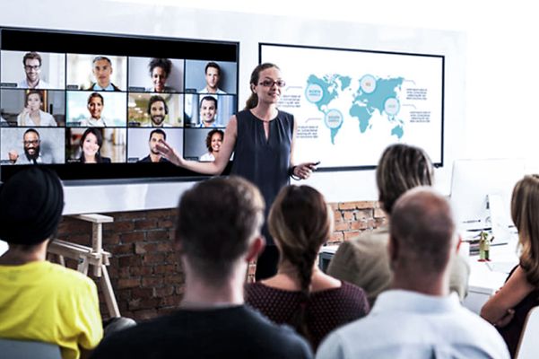 Hybrid meeting: A woman speaks to an audience on site and simultaneously to Zoom participants on a large screen.
