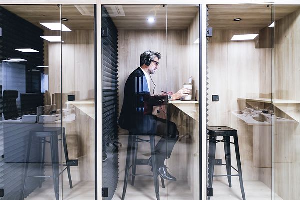 A manager in a soundproof booth, wearing headphones, is engrossed in work in a modern office.