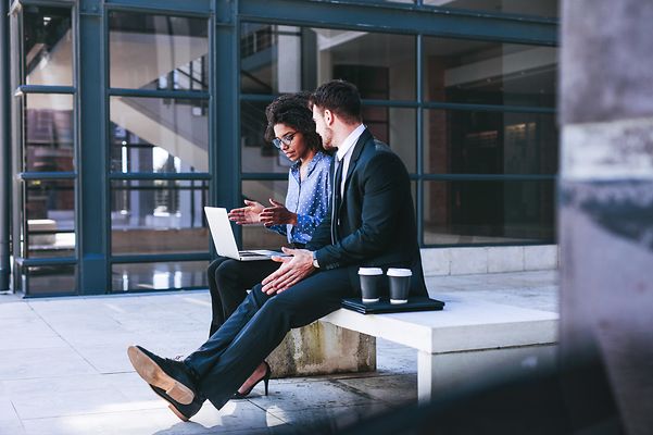 Zwei Kollegen sitzen vor einem Bürogebäude und schauen auf ein Notebook. Auf der Band neben ihnen stehen 2 Kaffee-to-Go Becher