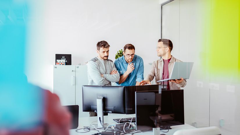 Three men are standing in front of a desk discussing suitable telephone systems.