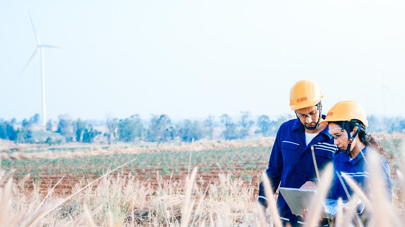 Two technicians in a field using Salesforce Energy & Utilities Cloud on a laptop, with wind turbines in the background