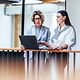 Two businesswomen are chatting whilst using a laptop.