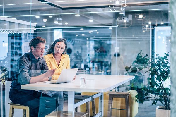 Two colleagues look at a project together on a laptop in a meeting room.