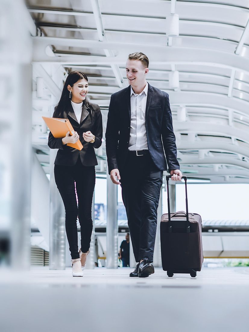 Two young businessmen walk past a building with their luggage.