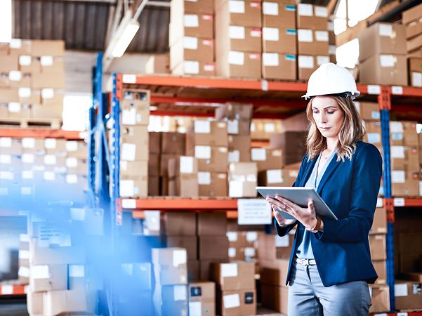 A young woman wearing a safety helmet stands in a warehouse and checks a list.