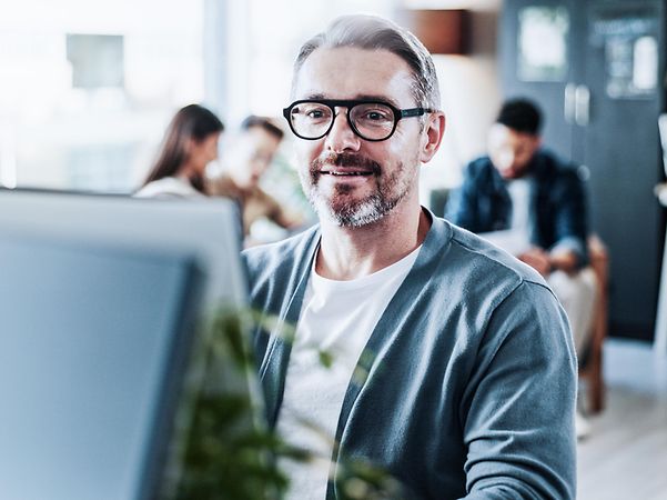 Man in office at computer, team meeting in background