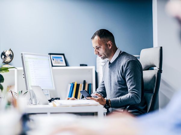A businessman sits in his office working with his laptop and smartphone.
