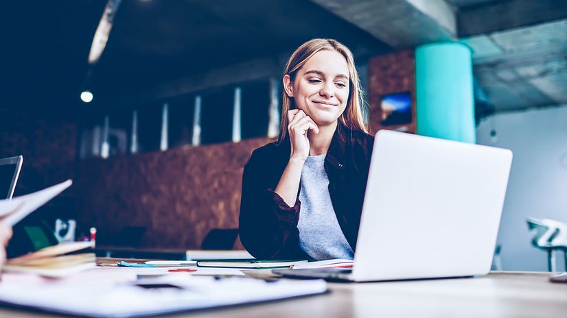 A woman sits at a table with documents in a modern office and looks at the screen.