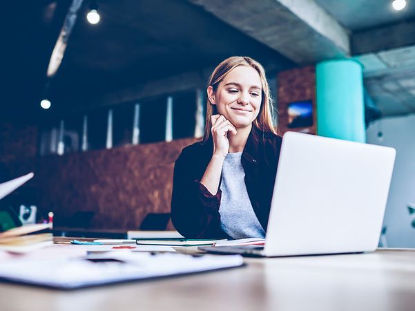 A woman sits at a table with documents in a modern office and looks at the screen.