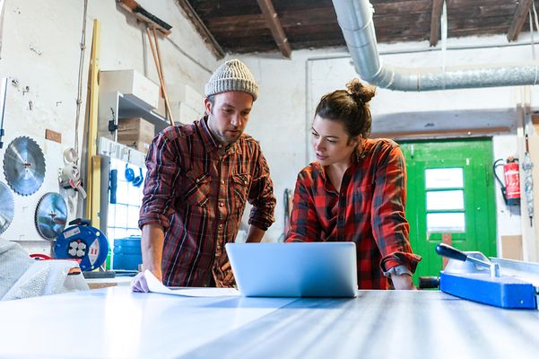 Handwerkerin und Handwerker mit Plan und Laptop bei der Diskussion in ihrer Werkstatt