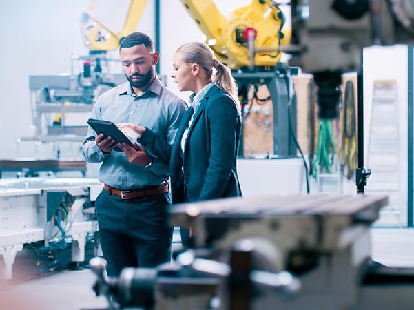 Two engineers are standing in a modern production facility, discussing something on a tablet.