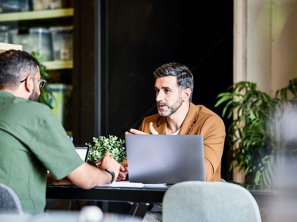 A man sits in front of his laptop and talks to another man.