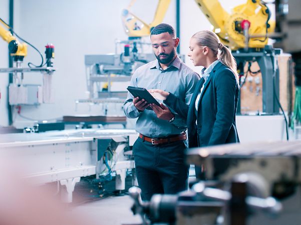Two engineers are standing in a modern production facility, discussing something on a tablet.