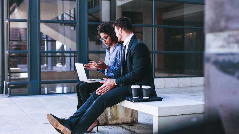 A woman and a man are sitting on a bench in front of a building, working on their mobile devices.