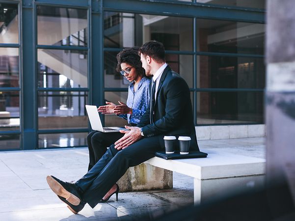 A woman and a man are sitting on a bench in front of a building, working on their mobile devices.