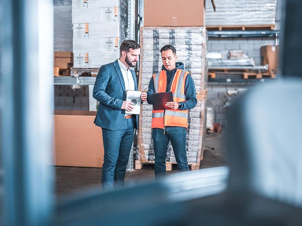 A man in business attire and a man in a high-visibility vest exchange information in a warehouse.