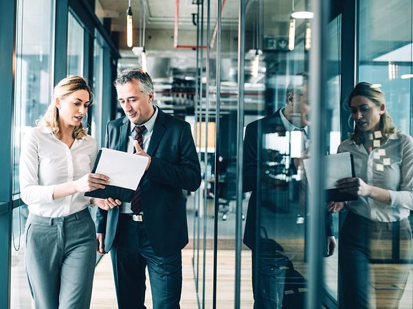 A man and a woman are walking down a corridor and looking at a tablet.