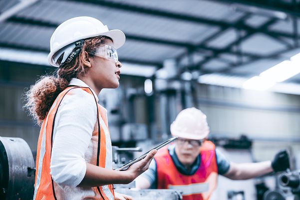 A woman is standing in a hall wearing a helmet and high-visibility vest, holding a tablet in her hand.