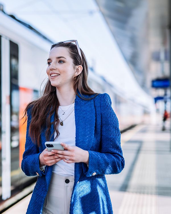 A woman stands smiling on the platform next to a train, holding a smartphone in her hand.