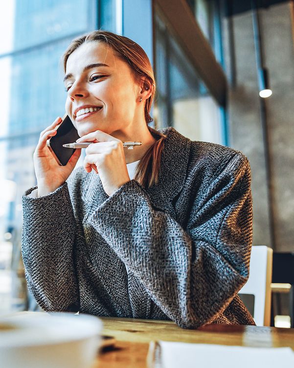Eine Frau sitzt in einem Kaffee, hat dank Local Listing eine gesuchte Rufnummer gefunden und telefoniert fröhlich.