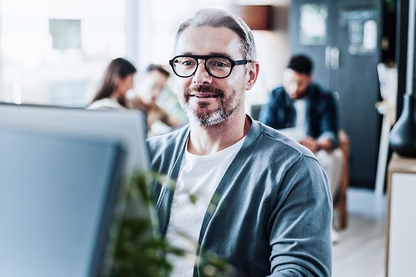 Man in office at computer, team meeting in background