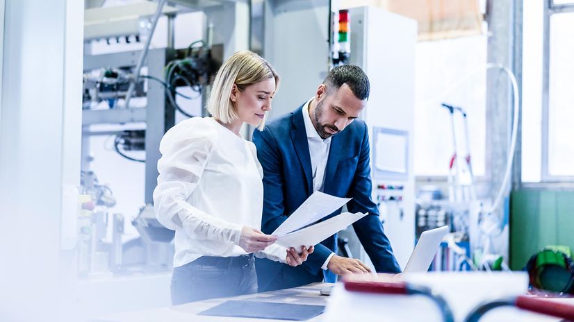 Businessman and young woman talking with papers and laptop in a factory