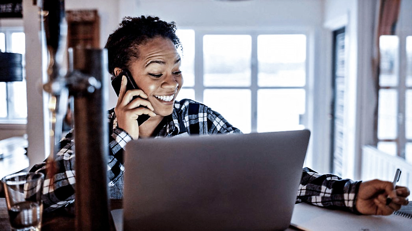 Woman sitting in front of a laptop, talking on her mobile phone and taking notes.