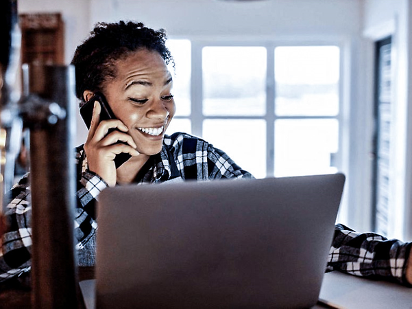 Woman sitting in front of a laptop, talking on her mobile phone and taking notes.