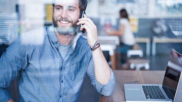 A man sits in front of his laptop with his mobile phone in his hand and smiles.