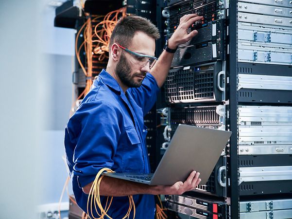A young man with a laptop is working with internet equipment and cables in the server room