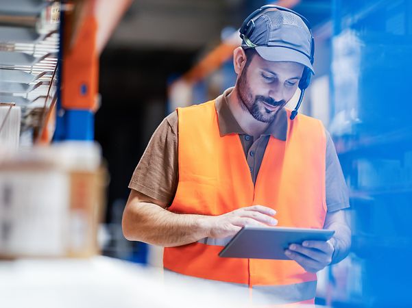 A man is standing in the warehouse looking at his tablet.