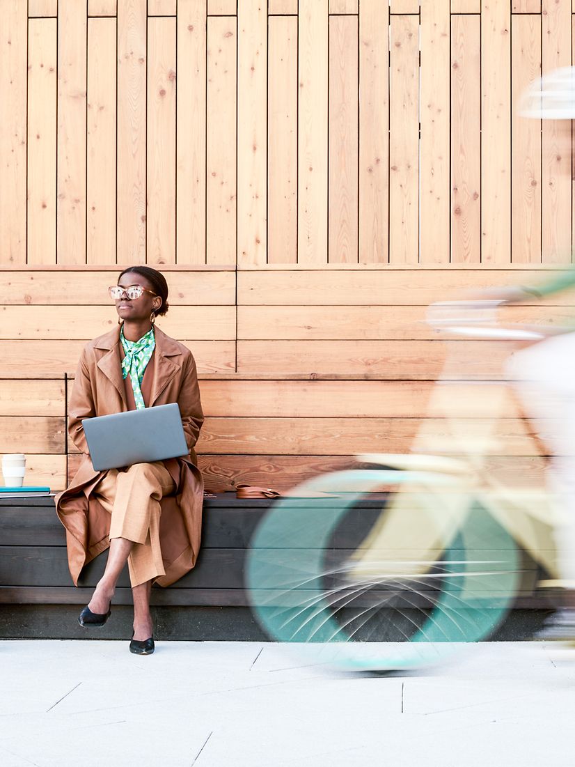 Woman sitting on a bench with a laptop, a cyclist rides through the foreground