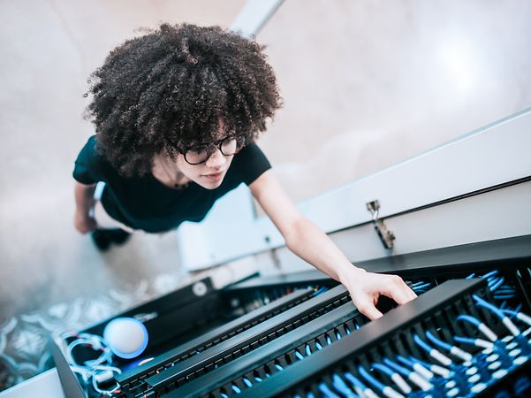 A young woman stands in front of a network server panel and checks a cable.