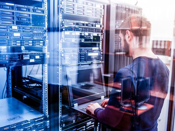 A young man holding a laptop stands in the server room and works on a server.