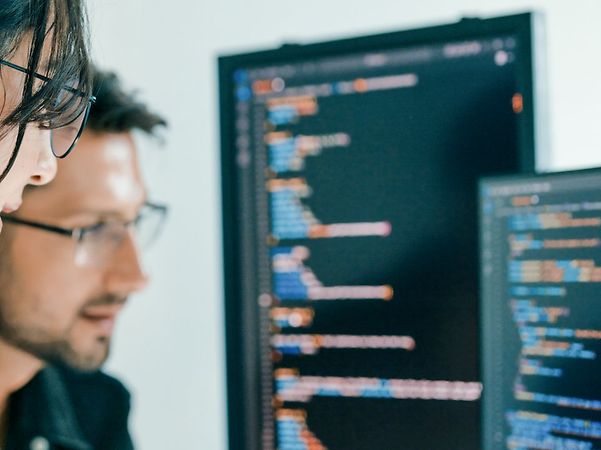 Zwei Personen analysieren Quellcode auf großen Monitoren, eine zeigt mit dem Finger auf den Bildschirm.